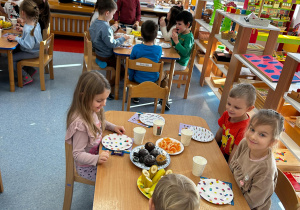 przedszkolaki siedzą przy stolikach i jedzą babeczki oraz owoce