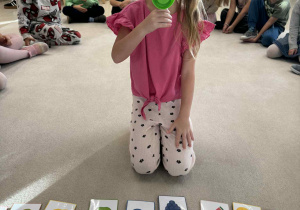 dziewczynka siedzi w środku koła i trzyma megafon zmiejący głos i wypowiada nazwe owocu w j.angielski