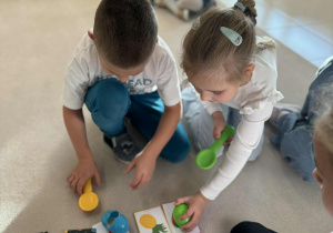 chłopiec i dziewczynka trzymają w dłoni platikowe łyzki z plasikowymi jajkami i kładą na odpowiedni owoc według polecenia nauczyciela