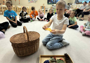 chłopiec siedzy w środku koła, przed nim jest taca z darami jesieni i koszyk a on trzyma żółty liść