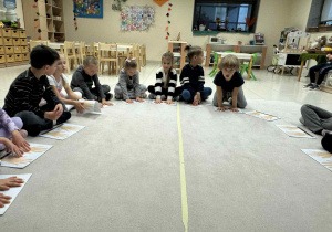 dzieci siedzą w kole i mają przed sobą kartki z dłoniami, w które uderzaja swoimi rękami wypowiadając przy tym nazwe owocu w j.angielskim