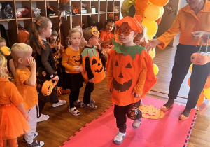 chłopiec w dyniowym stroju oraz dyniowych okularach przechodzi przez wybieg, za nim stoi pani w pomarańczowej bluzce.obok siedzą dzieci