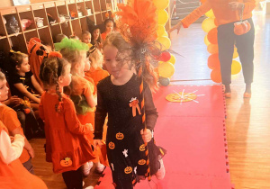 dziewczynka w czarno-pomarańczowym stroju, trzymająca ozdobioną miotłe, przechodzi po wybiegu