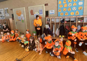 dzieci siedzą na podłodze w dyniowych strojach, za nimi stoi pani w pomarańczowej bluzce