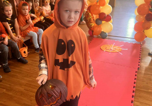 chłopiec w dyniowym stroju przechodzi po wybiegu i trzyma w dłoni lampion dyniowy