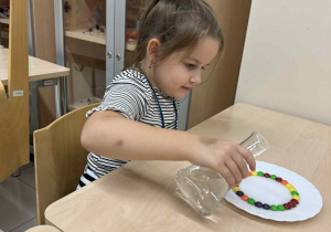 dzieczynka siedzi przy stoliku i nalewa wody na talerzyk z kolorowymi cukierkami