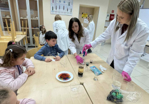 Pani nauczycielka Chemi i jej uczenica pokazują dzieciom eksperyment przy stoliku