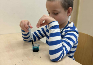 chłopiec siedzi przy stoliku i pipetą nelewa do pojemnika kolor zielony