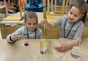 dwie dziewczynki siedzą przy stoliku trzymają pipety i wykonują eksperyment