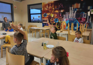 dzieci siedzą przy stoliki i słuchaja Pani od Chemi, jeden chłopiec podnosi rękę