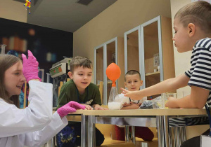 dzieci siedzą przy stoliki, obok nich jest uczenica i pokazuje im doświadczenie z balonem