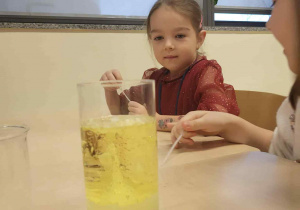 dziewczynka siedzi przy stoliku i patrzy na reakcje chemiczne w szkalnce