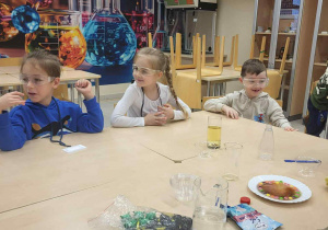 dwaj chłopcy i dziewczynka siedzą przy stoliku i mają na sobie okulary ochronne