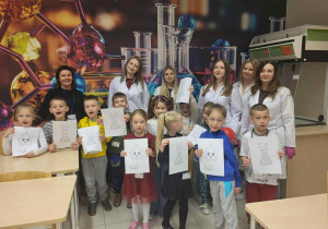 dzieci trzymając kolorwanki wraz z Paniami i uczenicami pozują do wspólnego zdjęcia w klasie podczas zajęć z Chemi