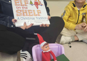 Pani siedzi trzymając ksiązke i opowiada historie o "Elf on the shelf" obok siedzi chłopiec w żółtej bluzce