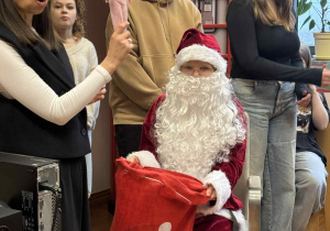 mikołaj siedzi na krześle i trzyma worek z prezentami a obok stoi Pani od angielskiego i uczniowie