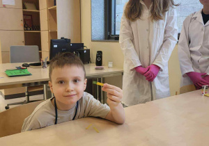 chłopiec siedzi przy stoliku, trzymając zabarwiony papierek za nim stoją uczenice w białych fartuchach