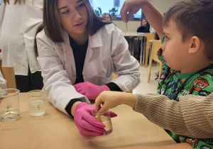 uczenica licem rozdaje dzieciom papierowe papierki, obok niej siedzi chłopiec z ręką w górze