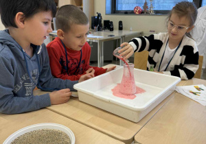 dzieci siedzą przy stoliku i dziewczynka nalewa ocet do naczynia aby powstał wulkan