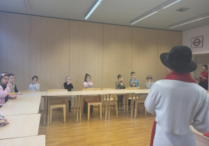 dzieci stoją przed stolikami i śpiewają piosenkę z uczenicą liceum w jęzku angielskim