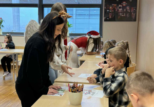 dzieci siedzą przy stolikach a nad nimi stoją uczenice liceum, które im pomagają w zadaniach w języku angielskim