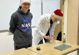 dwaj uczniowe technikum w mikołajowych czapach stoją za stołem i pokazyją eksperyment