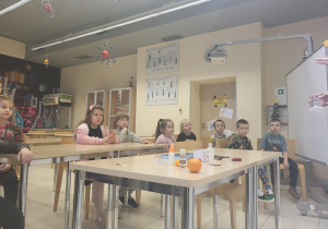 dzieci siedzą przy stolikach w półkolu,obserwujac i słuchajac podczas zajęć z fizyki