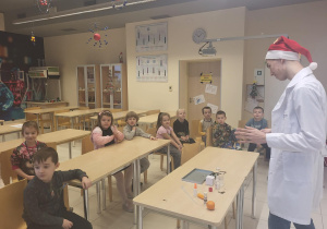 dzieci siedzą przy stolikach i obserwują eksperymenty robione przez ucznia