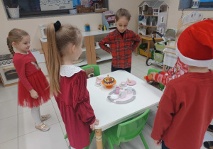 dzieci stoją nad stolikiem, na którym jest drewniany torcik, talerze i zastawa do herbaty