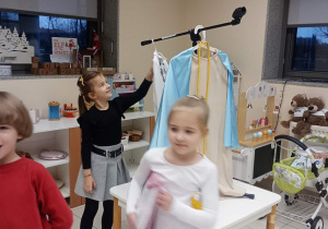 uśmiechnieta dziewczynka patrzy na statyw od mikforonu, na którym wisi strój i jest ustawiony na stole