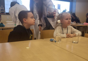chłopiec obserwuje doświadczenie wykonane w słoiku przez uczenice, obok nich siedzi dziewczynka