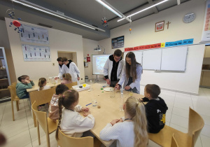 dzieci siedzą przy stoliku i obserwują uczniów, wykonujących eksperyment chemiczny