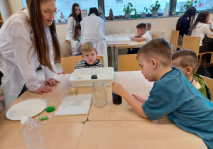chłopcy siedzą przy stoliku przy nich stoi uczenica i wykonują doświadczenie