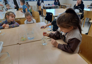 dziewczynka wykonuje doświadczenie,obok niej siedzą inne dziewczynki przypatrując się jej