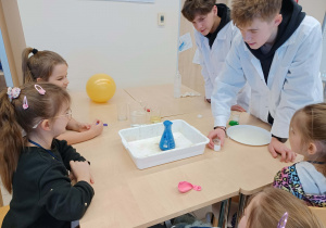 dziewczynki siedzą przy stoliku i wykonują doświadczenie chemiczne a przy nich stoją uczniowe