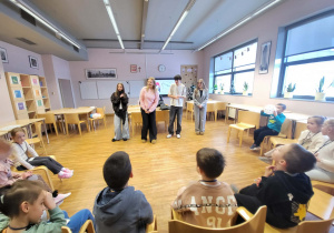 dzieci siedzą w półkolu a przed nimi stoją uczniowe