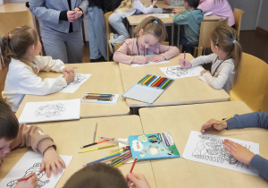dzieci siedzą przy stoliku i kolorują, przy nich stoi uczenica