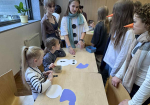 dwie dziewczynki siedzą przy stoliku i robią papierowe bałwanki a przy nich stoją uczenice liceum