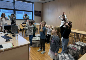 dzieci stoja w półkolu z nauczycielka angielskiego,pokazując i śpiewająć piosenke po angielsku