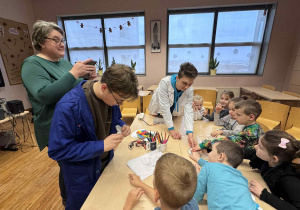 dzieci siedzą przy stoliku i eksperymentują z ciepłem i zimnem a nad nimi stoją uczniowie i pani od fizyki