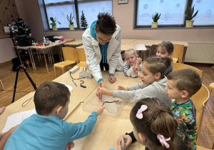 dzieci siedzą przy stoliku i jeden z pomocą ucznia technikum próbuje przeciąć lód