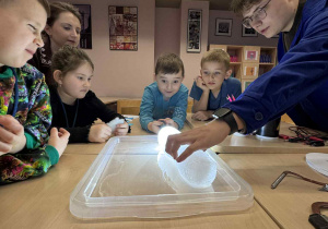 dzieci siedzą przy stoliku i obserwują jak jeden chłopiec podświetla lód