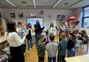 dzieczi stoją w kole z uczenicami liceum i panią od angielskiego, śpiewając i pokazując
