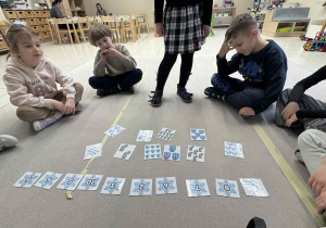 dzieci siedzą w kole i jedną dziewczynka stoji i wykonuję zadanie z liczeniem ubrań po angielsku i dopasowaniem do odpowiedniej liczby