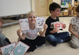 uśmiechnieta dziewczynka trzyma dwa obrazki z pączkami, obok niech siedzi chłopiec, który też ma parę obrazków z pączkami