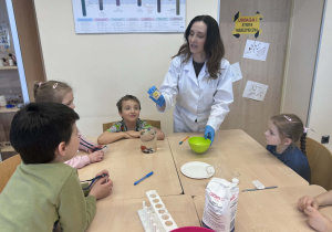 dzieci siedzą w ławce i obserwują Pania, która wykonuję eksperyment z lodem