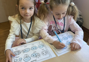 dwie dziewczynki siedzą przy stoliku i koloruja obrazek z pogodą