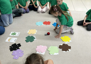 dzieci siedzą w kole na środku jest dziewczynka, która kręci zabawką "wskazujacy paluszek"i wykonuję zadanie z kolorowymi kończynkami