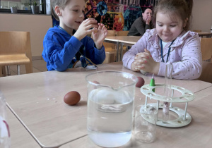 chłopiec i dziewczynka siedzą przy stoliku i próbuja zgnieść jajko zawinięte w folię aluminiową