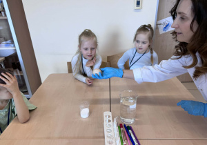 od chemii trzyma jajko bez skorupki pokazując je dzieciom siedzącym przy stoliku